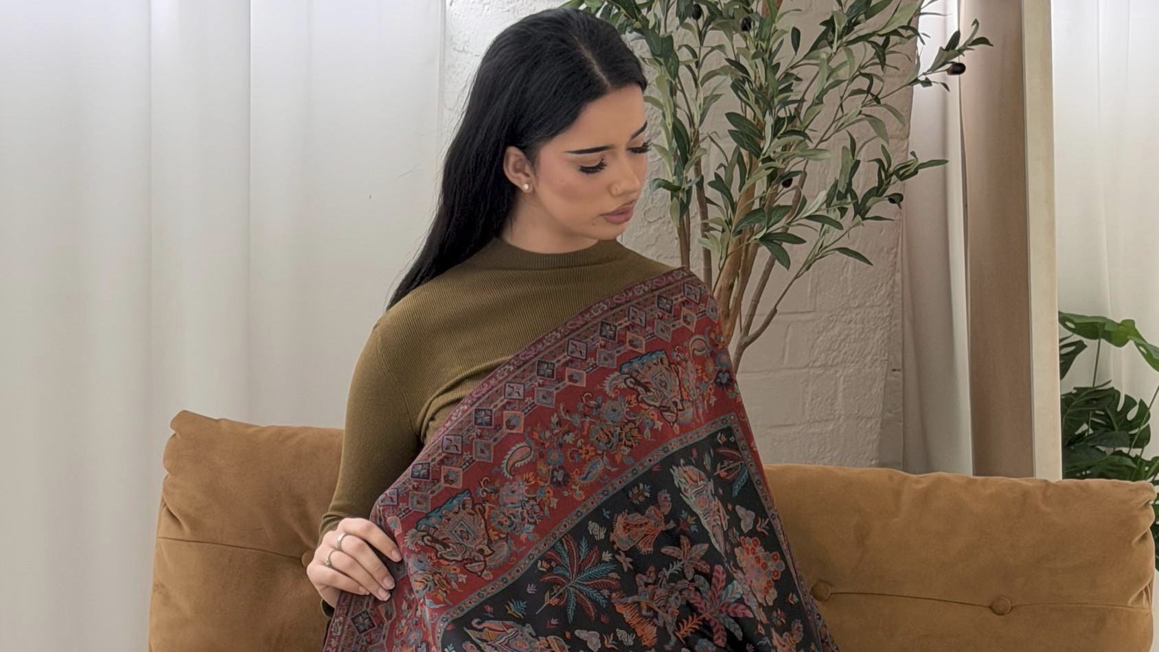 Woman holding a black patterned pashmina shawl scarf in a room with a couch and plants.