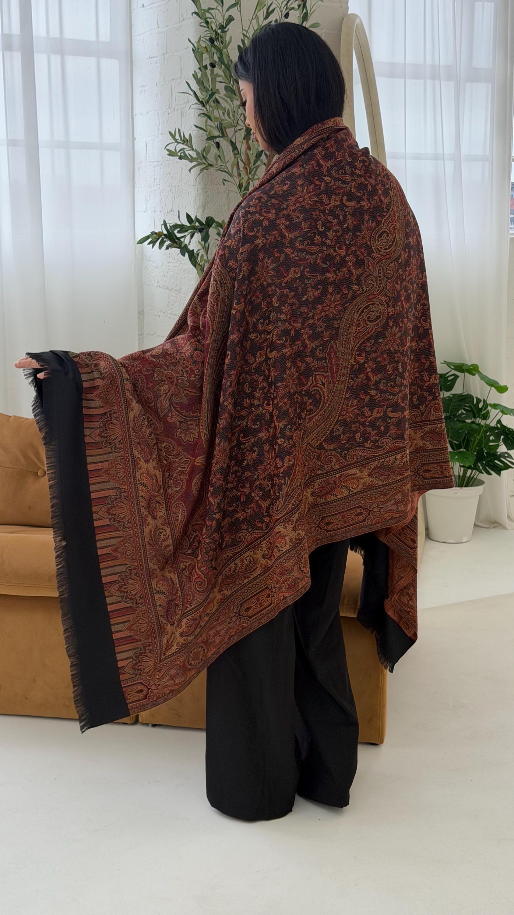 Person draped in a patterned brown and black shawl in a room with white curtains and plants.