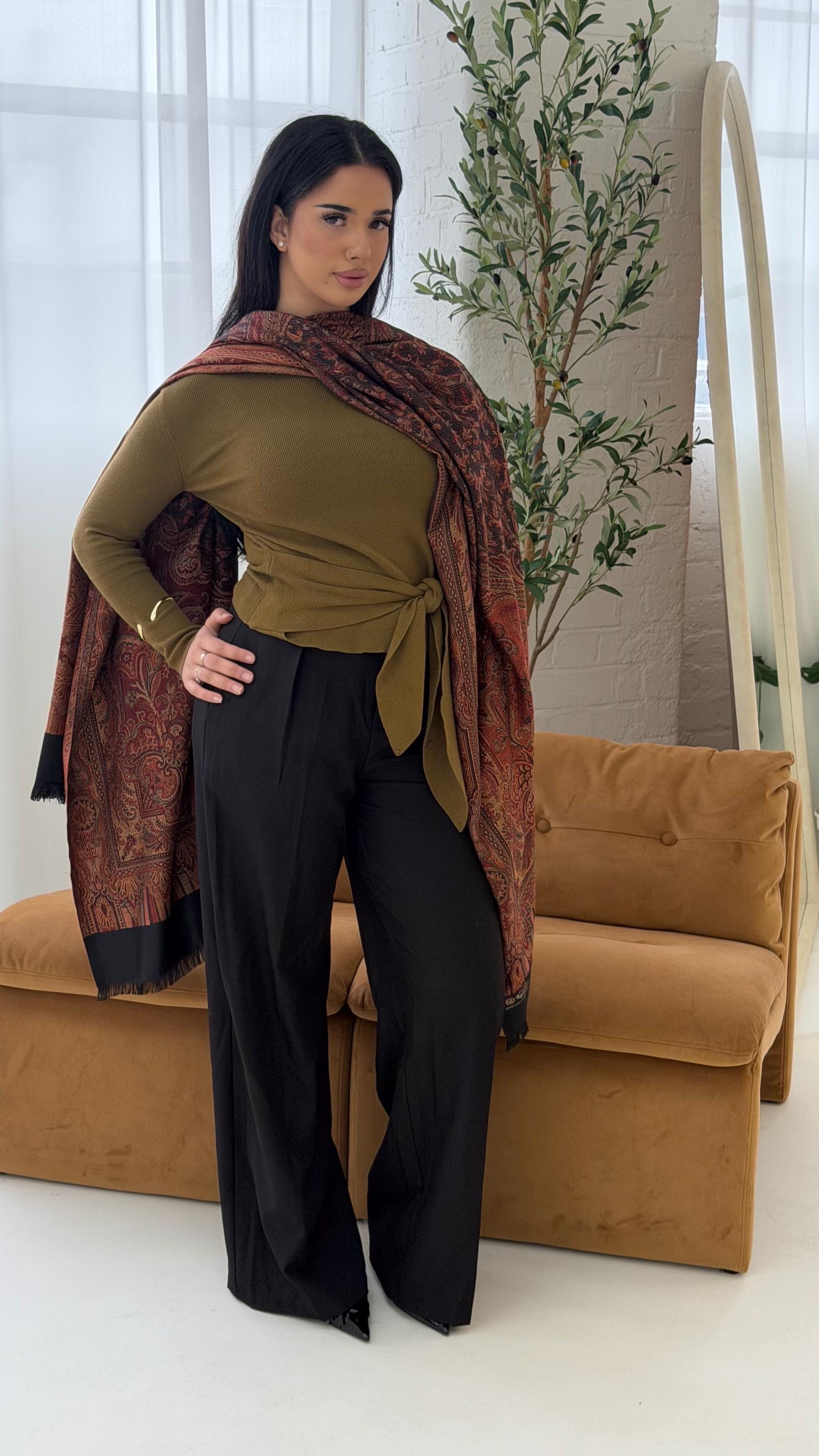 Woman in green top and black pants with a black pashmina patterned shawl standing in a room with a brown couch and plant.