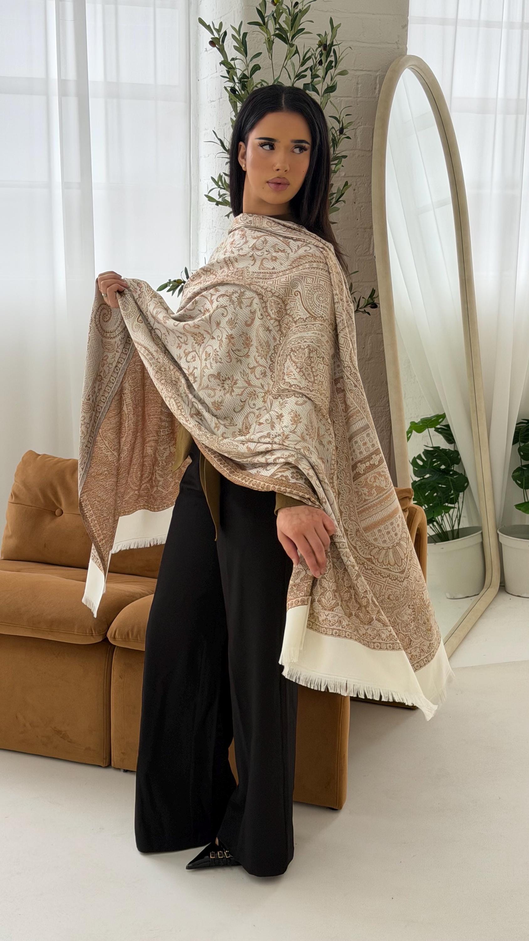 Woman holding a cream patterned shawl in a room with a couch and plants.