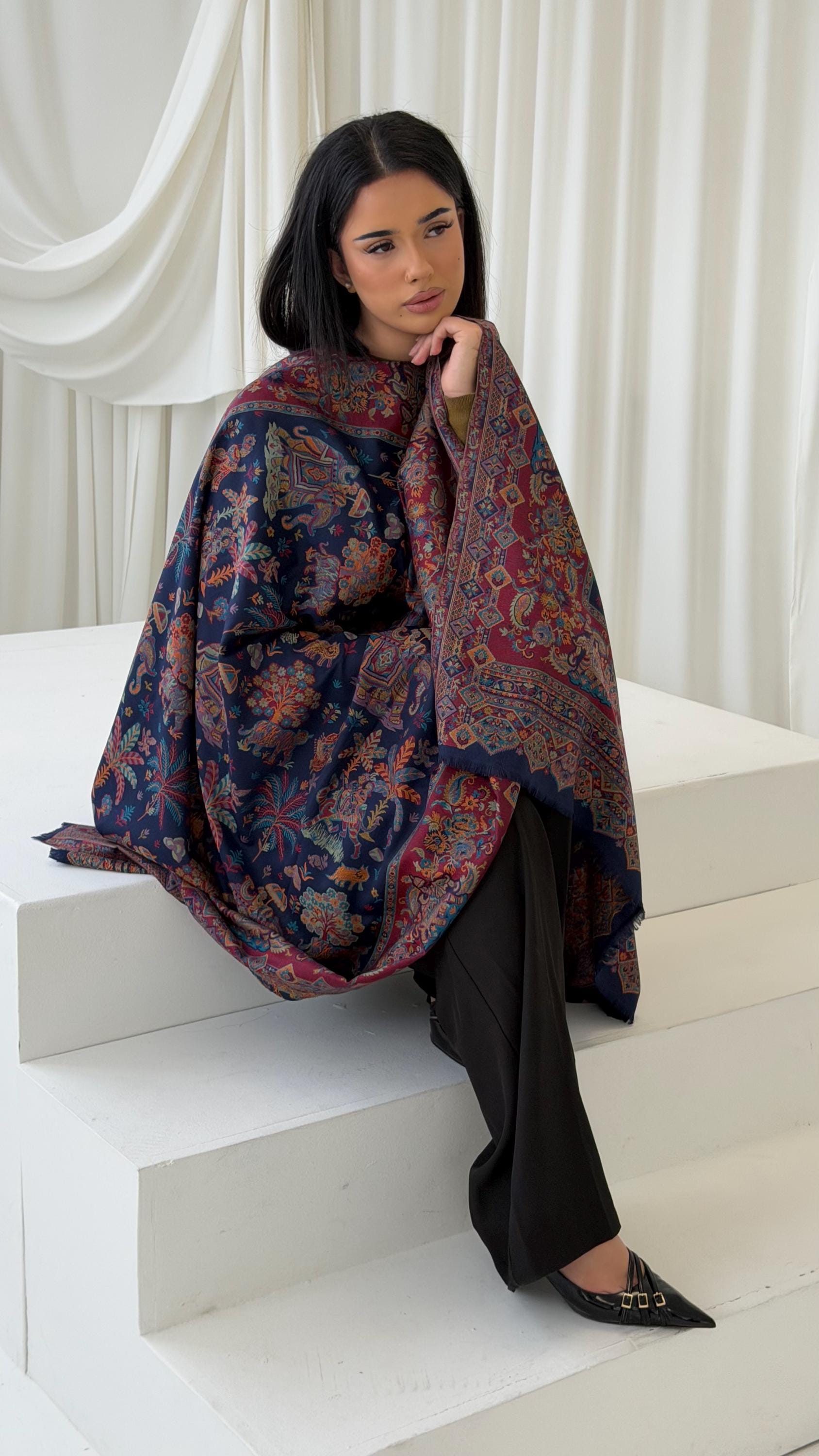 Woman sitting on white steps wearing a blue patterned pashmina shawl scarf against a white curtain background