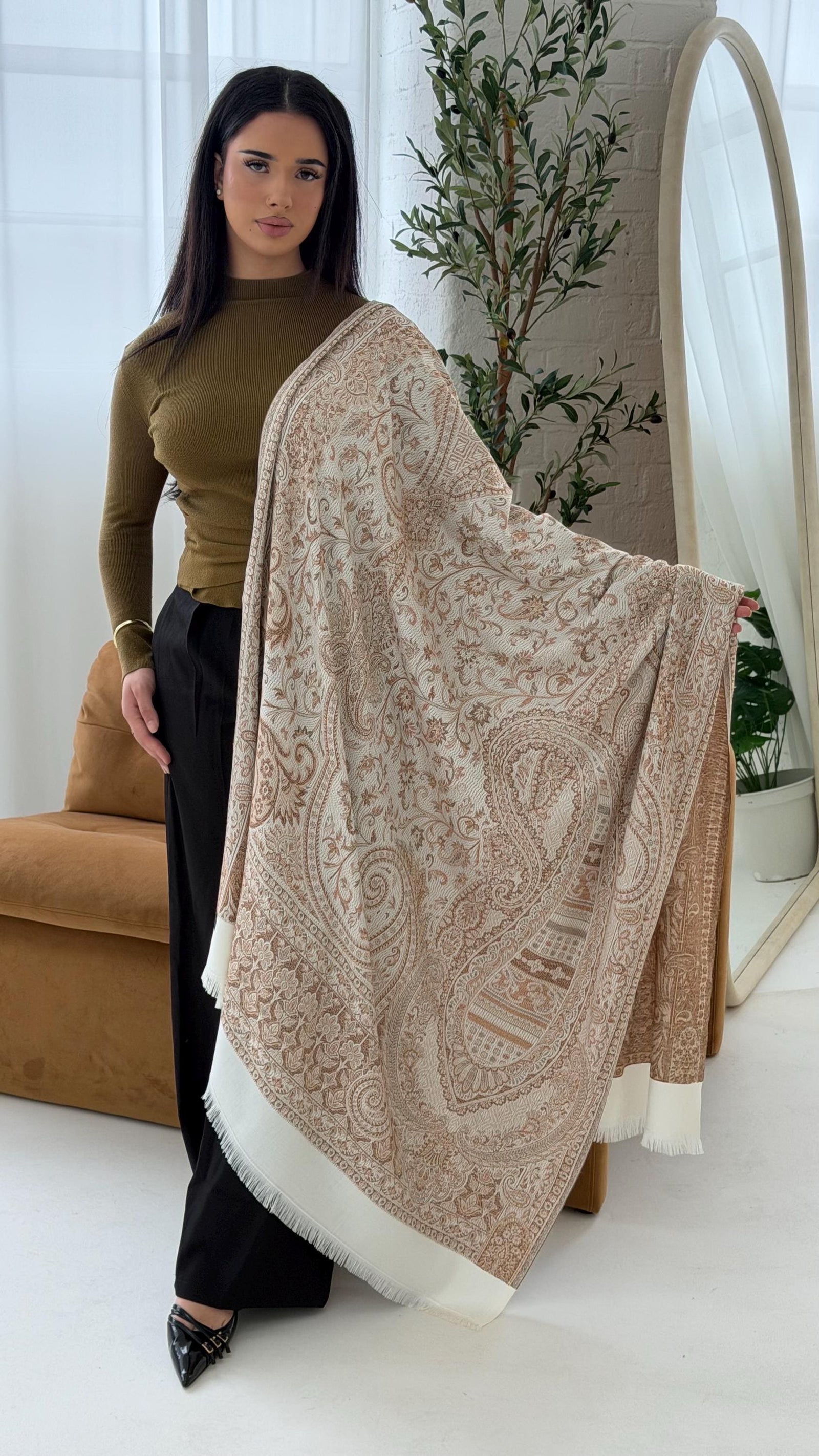 Woman holding a large beige patterned scarf in a bright room with plants.